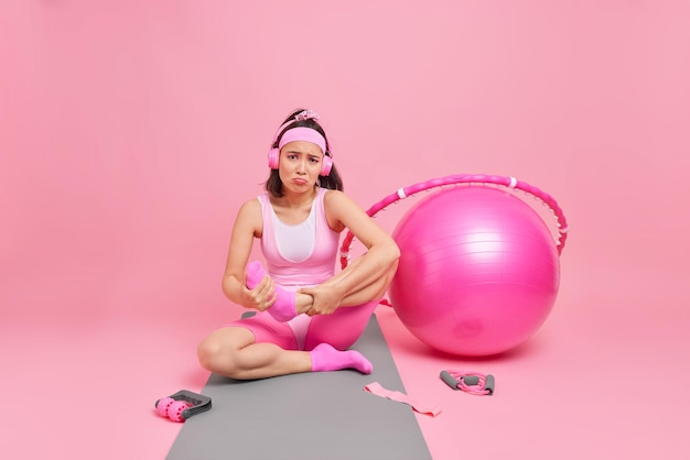 Junge frustrierte sportliche frau leidet unter schmerzen im bein, nachdem sie auf einer fitnessmatte trainiert hat, um sich zu bewegen, hört musik über kopfhörer in aktivkleidung und verwendet sportgeräte, die an einer rosa wand isoliert sind