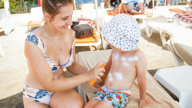 Junge fürsorgliche Mutter, die ihrem kleinen Kleinkindsohn am Meeresstrand Sonnencreme aufträgt.