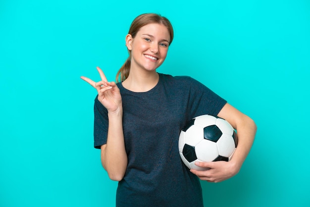 Junge Fußballspielerin isoliert auf blauem Hintergrund lächelnd und Victory-Zeichen zeigend