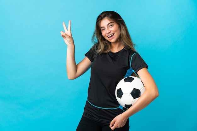 Junge Fußballspielerin lokalisiert auf der blauen Wand lächelnd und siegreiches Zeichen zeigend