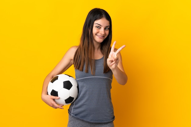 Junge Fußballspielerin lokalisiert auf gelbe Wand lächelnd und Siegeszeichen zeigend
