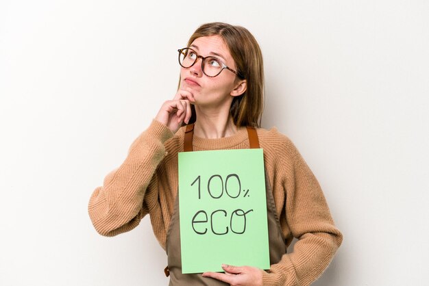Junge Gärtnerin, die ein 100-Öko-Plakat isoliert auf weißem Hintergrund hält und mit zweifelndem und skeptischem Ausdruck zur Seite schaut