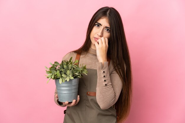 Junge Gärtnerin, die eine Pflanze lokalisiert auf rosa Wand hält, die Zweifel hat