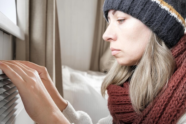 Junge gefrorene Frau, die einen Pullover trägt, der für Winterkälte mit Schockausdruck im Gesicht zittert und einfriert Mädchen wärmt die Hände mit Atem über der elektrischen Heizung auf