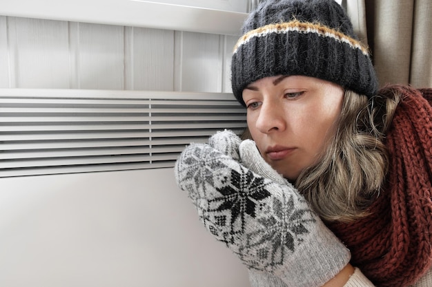 Junge gefrorene Frau, die einen Pullover trägt, der für Winterkälte mit Schockausdruck im Gesicht zittert und einfriert Mädchen wärmt die Hände mit Atem über der elektrischen Heizung auf