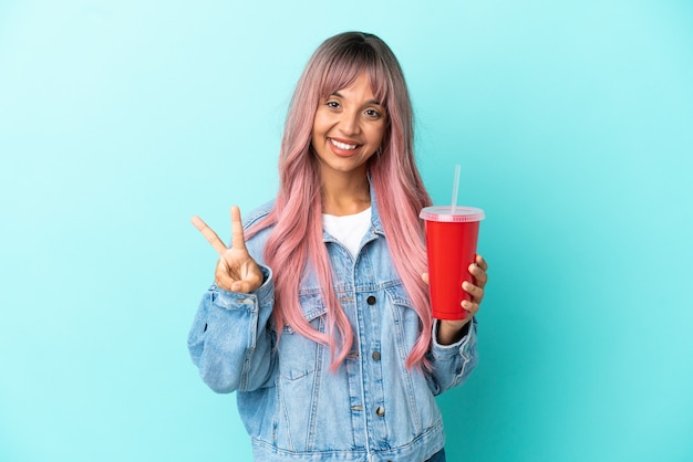 Junge gemischte Frau, die ein frisches Getränk trinkt, isoliert auf blauem Hintergrund, lächelt und zeigt Siegeszeichen