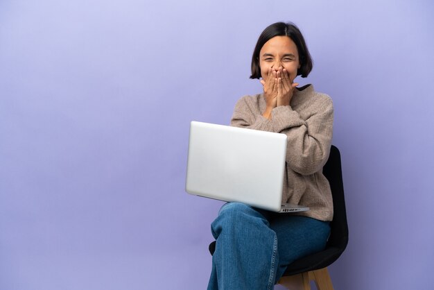 Junge gemischte frau sitzt auf einem stuhl mit laptop isoliert auf lila hintergrund glücklich und lächelnd den mund mit den händen bedeckend