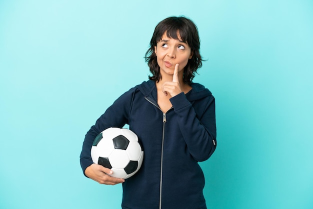 Junge gemischte Fußballspielerin isoliert auf blauem Hintergrund, die Zweifel hat, während sie nach oben schaut