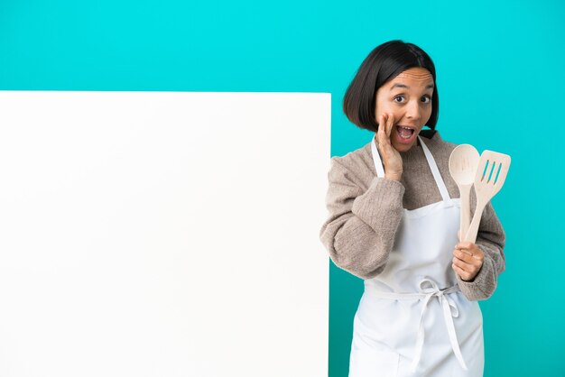 Junge gemischte kochfrau mit einem großen plakat einzeln auf blauem hintergrund, das auf die seite zeigt, um ein produkt zu präsentieren und etwas zu flüstern