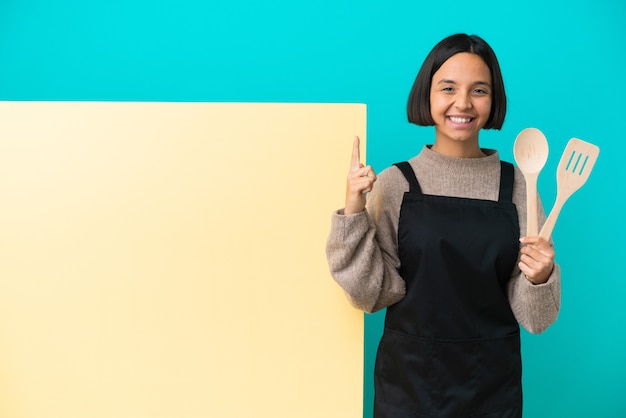 Junge gemischte Kochfrau mit einem großen Plakat isoliert auf blauem Hintergrund, das auf eine großartige Idee hinweist