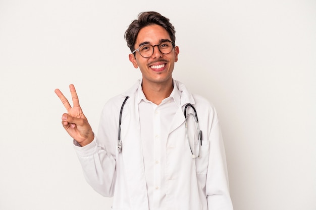 Junge gemischte Rasse Arzt Mann isoliert auf weißem Hintergrund freudig und sorglos zeigt ein Friedenssymbol mit den Fingern.