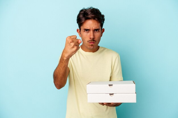Junge gemischte Rasse Mann hält Pizzas isoliert auf blauem Hintergrund mit Faust zur Kamera, aggressiven Gesichtsausdruck.