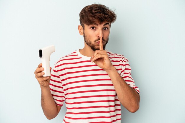 Junge gemischte Rasse Mann hält Thermometer isoliert auf blauem Hintergrund hält ein Geheimnis oder bittet um Stille.