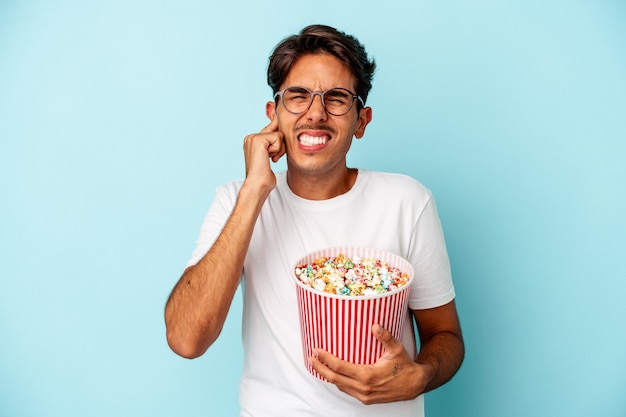 Junge gemischte Rasse Mann isst Popcorn isoliert auf blauem Hintergrund für Ohren mit Händen.