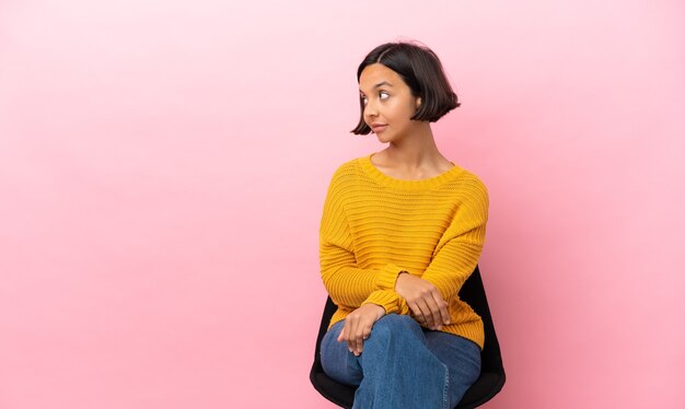 Junge gemischte Rassenfrau, die auf einem Stuhl lokalisiert auf rosa Hintergrund in der hinteren Position sitzt und Seite schaut
