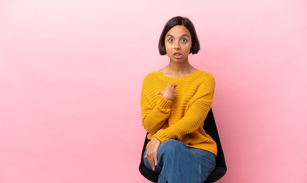 Junge gemischte Rassenfrau, die auf einem Stuhl lokalisiert auf rosa Hintergrund sitzt und auf sich selbst zeigt