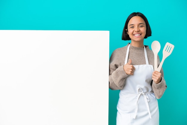 Junge gemischte Rassenkochfrau mit einem großen Plakat lokalisiert auf blauem Hintergrund, der eine Daumen hoch Geste gibt