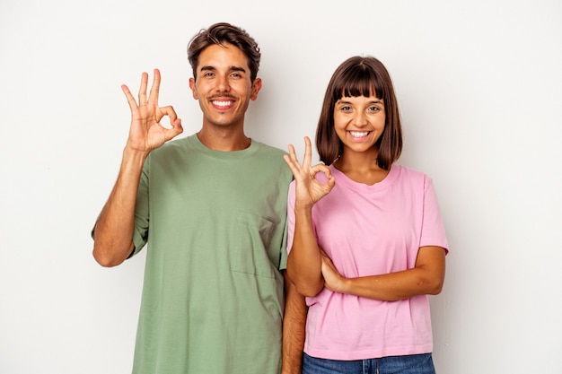 Foto junge gemischte rassenpaare, die auf weißem hintergrund isoliert sind, zwinkert ein auge und hält eine okaygeste mit der hand.