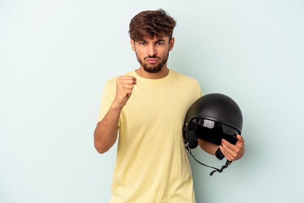 Junge gemischte Rennen Mann mit Motorradhelm isoliert auf blauem Hintergrund mit Faust zur Kamera, aggressiven Gesichtsausdruck.