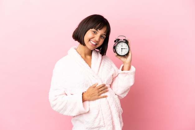 Junge gemischte schwangere Frau isoliert auf rosa Hintergrund im Pyjama und hält Uhr mit glücklichem Ausdruck