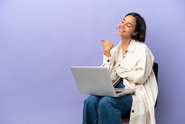 Junge gemischtrassige Frau, die auf einem Stuhl mit Laptop sitzt, isoliert auf violettem Hintergrund, der auf die Seite zeigt, um ein Produkt zu präsentieren?