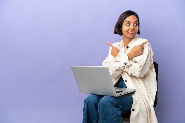 Junge gemischtrassige Frau, die auf einem Stuhl mit Laptop sitzt, isoliert auf violettem Hintergrund, der auf die Seitenwände zeigt, die Zweifel haben?