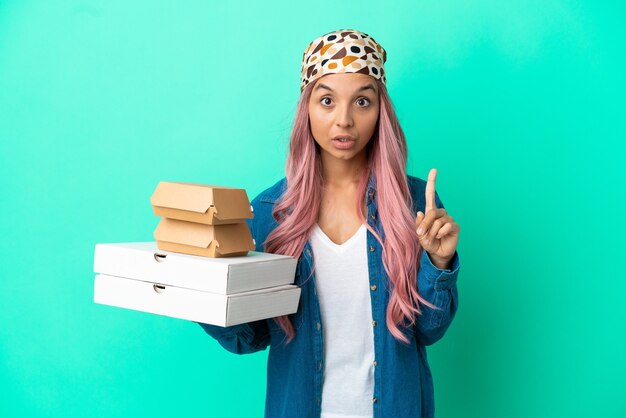 Junge gemischtrassige Frau, die Pizza und Burger einzeln auf grünem Hintergrund hält, die beabsichtigt, die Lösung zu realisieren, während sie einen Finger nach oben hebt