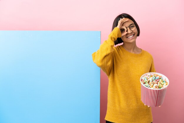 Foto junge gemischtrassige frau, die popcorn mit einem großen banner über isoliertem hintergrund hält, das ein ok-zeichen mit den fingern zeigt