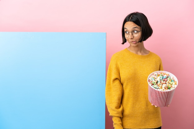 Junge gemischtrassige Frau, die Popcorn mit einem großen Banner über isoliertem Hintergrund hält und beim Nachschlagen Zweifel hat