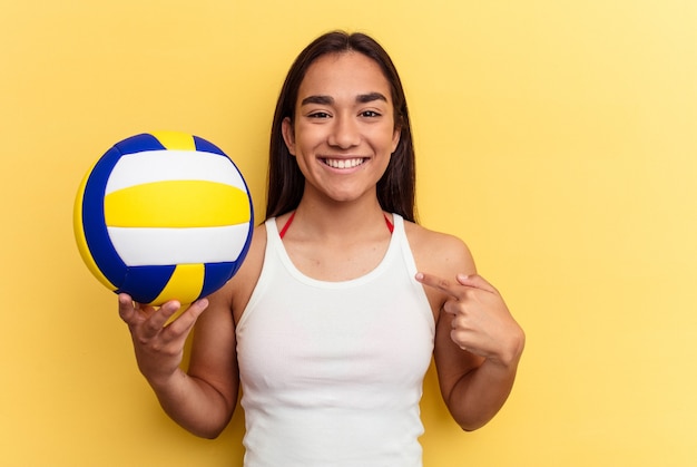 Junge gemischtrassige Frau, die Volleyball am Strand spielt, einzeln auf gelbem Hintergrund, Person, die mit der Hand auf einen Hemdkopierraum zeigt, stolz und selbstbewusst