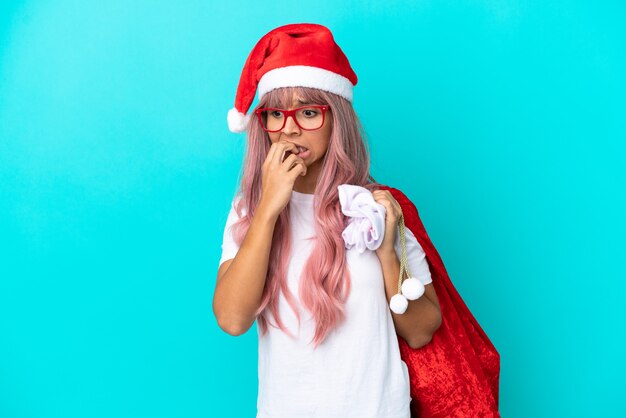 Junge gemischtrassige Frau, die zu Weihnachten Geschenke verteilt auf blauem Hintergrund mit Zweifeln isoliert