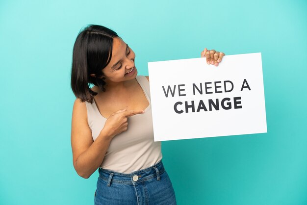 Junge gemischtrassige Frau isoliert auf blauem Hintergrund, die ein Plakat mit dem Text We Need a Change hält und darauf zeigt