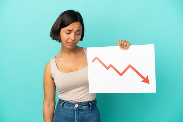 Junge gemischtrassige Frau isoliert auf blauem Hintergrund, die ein Schild mit einem abnehmenden Statistikpfeilsymbol mit traurigem Ausdruck hält