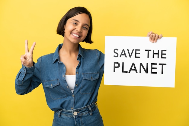 Junge gemischtrassige Frau isoliert auf gelbem Hintergrund, die ein Plakat mit dem Text Save the Planet hält und einen Sieg feiert