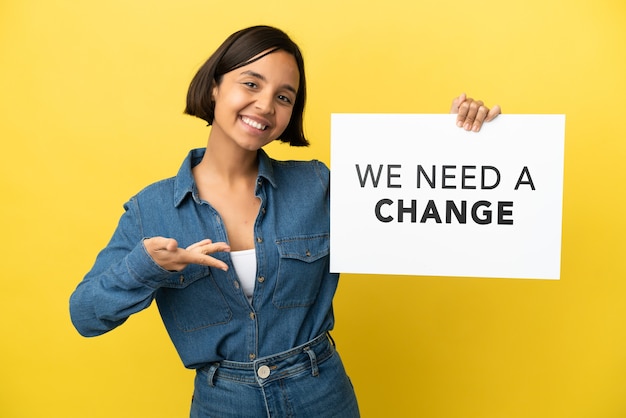Junge gemischtrassige Frau isoliert auf gelbem Hintergrund, die ein Plakat mit dem Text We Need a Change hält und darauf zeigt