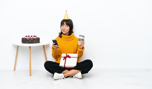 Junge gemischtrassige Frau mit einem Geschenk auf dem Boden sitzend isoliert auf weißem Hintergrund mit Kaffee zum Mitnehmen und einem Handy, während sie etwas denkt