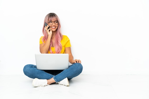 Junge gemischtrassige Frau mit einem Laptop mit rosa Haaren, die auf dem Boden sitzt, isoliert auf weißem Hintergrund, der ein Gespräch mit dem Mobiltelefon führt