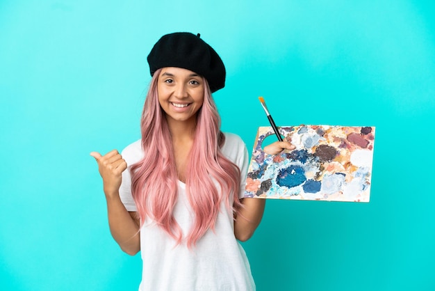 Junge gemischtrassige Frau mit rosa Haaren, die eine auf blauem Hintergrund isolierte Palette hält, die auf die Seite zeigt, um ein Produkt zu präsentieren