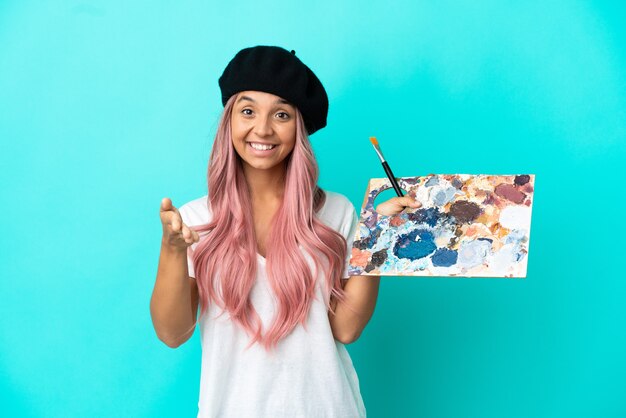 Junge gemischtrassige Frau mit rosa Haaren, die eine auf blauem Hintergrund isolierte Palette hält, die Hände schüttelt, um ein gutes Geschäft abzuschließen