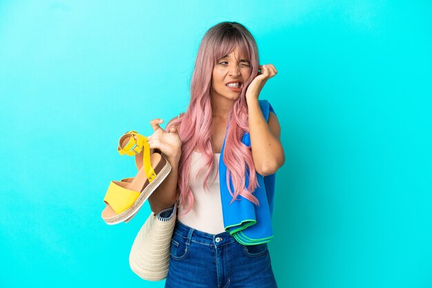 Junge gemischtrassige Frau mit rosa Haaren, die Sommersandalen auf blauem Hintergrund isoliert hält, frustriert und die Ohren bedeckend