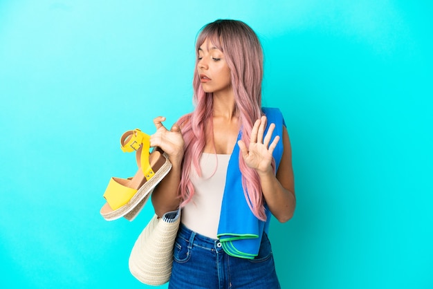 Junge gemischtrassige Frau mit rosa Haaren, die Sommersandalen isoliert auf blauem Hintergrund hält und Stoppgeste macht und enttäuscht