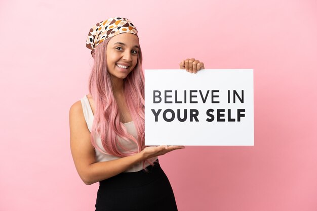 Junge gemischtrassige Frau mit rosa Haaren isoliert auf rosa Hintergrund, die ein Plakat mit dem Text Believe In Your Self hält und darauf zeigt
