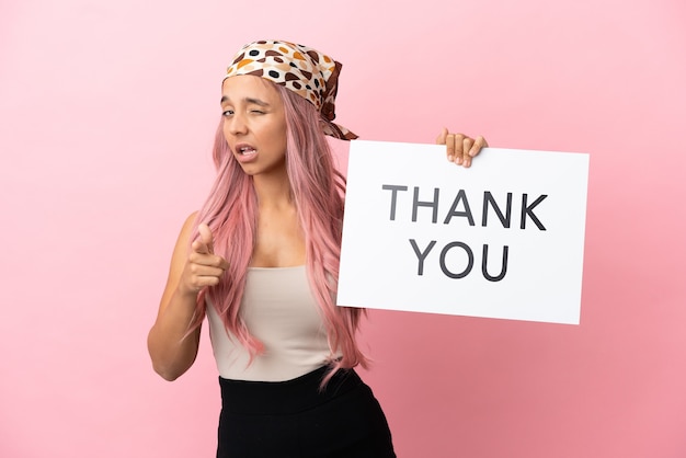 Junge gemischtrassige Frau mit rosa Haaren isoliert auf rosa Hintergrund, die ein Plakat mit dem Text DANKE hält und nach vorne zeigt