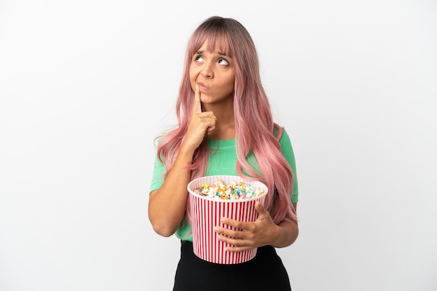 Junge gemischtrassige Frau mit rosa Haaren isst Popcorn isoliert auf weißem Hintergrund und hat beim Nachschlagen Zweifel