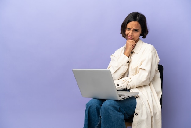 Junge gemischtrassige Frau sitzt auf einem Stuhl mit Laptop isoliert auf lila Hintergrund und hat Zweifel