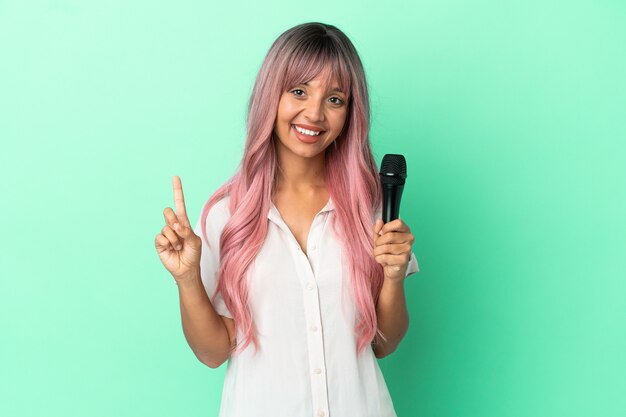 Junge gemischtrassige Sängerin mit rosa Haaren einzeln auf grünem Hintergrund, die einen Finger im Zeichen des Besten zeigen und heben