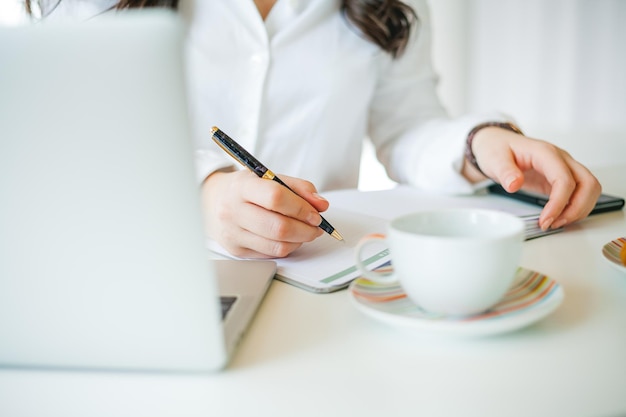 Junge Geschäftsfrau arbeitet am Computer Eine schöne dunkelhaarige Frau in einem weißen Hemd arbeitet online in einem hellen Büro Kaffee trinken und ein Croissant essen