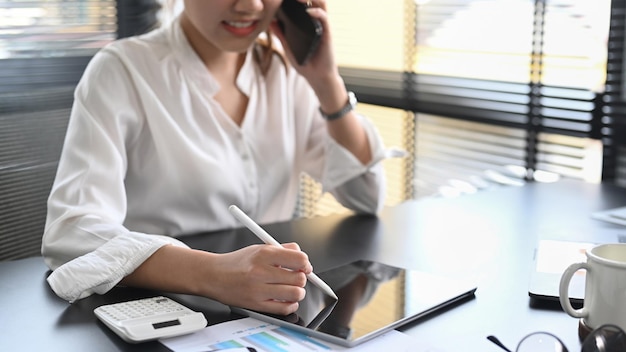 Junge Geschäftsfrau, die ein Handygespräch führt und ein digitales Tablet im modernen Büro verwendet