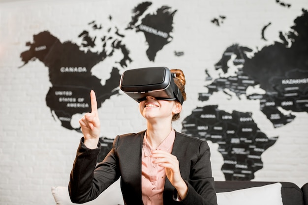 Foto junge geschäftsfrau, die eine virtual-reality-brille versucht, die drinnen mit weltkarte im hintergrund sitzt