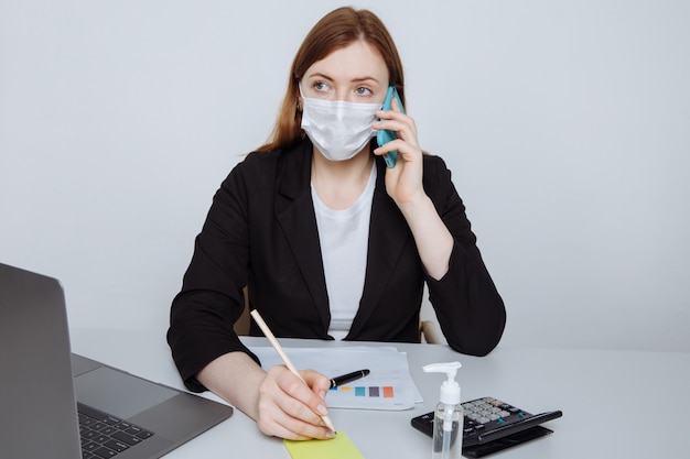 Junge Geschäftsfrau, die Gesichtsmaske spricht, die auf Smartphone sitzt, das am Arbeitsplatzschreibtisch sitzt